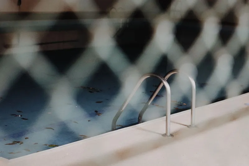 Piscina descuidada con agua oscura y hojas vista a través de una valla metálica